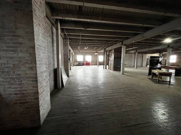 A wide, empty industrial loft room with dark, worn wooden floors and a rustic exposed brick wall on the left. The ceiling has exposed wooden beams. Several small windows are visible on the far wall, letting in light. A few pieces of furniture are scattered throughout the space.