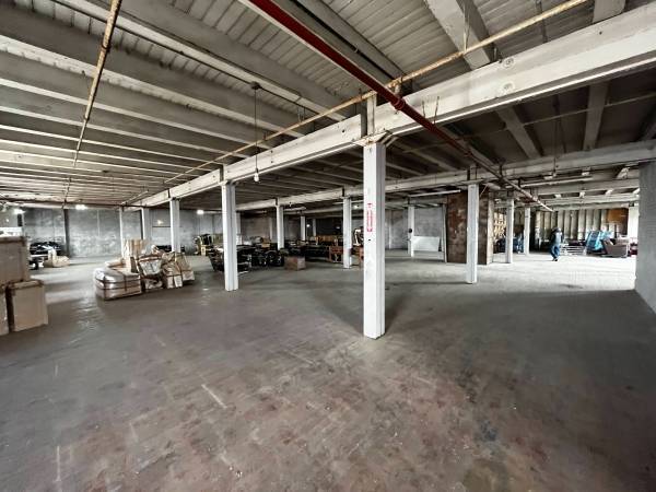 A wide shot of a large, empty industrial room with a gray concrete floor and numerous white metal support columns. The ceiling is high and shows exposed beams and pipes. Stacks of cardboard boxes and other materials are scattered around the room.