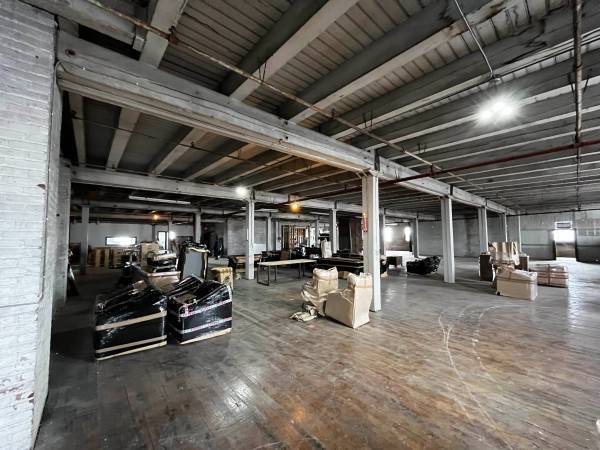 A wide-angle shot of a large, unfinished industrial room. The floor is a mix of dark wood and concrete. White metal beams and supports run across the ceiling and are held up by several white pillars. The space is largely empty, though some pieces of furniture wrapped in black plastic and several cardboard boxes are visible.
