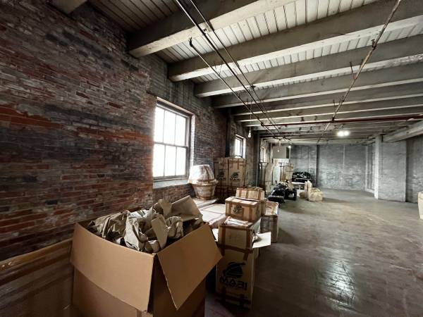 An interior shot of an industrial room with a brick wall on the left and a wood-paneled ceiling with exposed beams. A large open cardboard box filled with crumpled paper is in the foreground. Several other boxes and items are stacked along the wall beneath two windows.