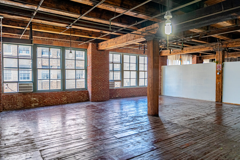 A sunlit, empty industrial loft space with exposed brick walls, a wooden beamed ceiling, and original hardwood floors. Large, multi-pane windows line the far wall, letting in natural light. In the foreground, a single thick wooden pillar supports the ceiling. The floor is aged and has visible wear and tear.