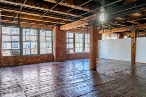 A sunlit, empty industrial loft space with exposed brick walls, a wooden beamed ceiling, and original hardwood floors. Large, multi-pane windows line the far wall, letting in natural light. In the foreground, a single thick wooden pillar supports the ceiling. The floor is aged and has visible wear and tear.