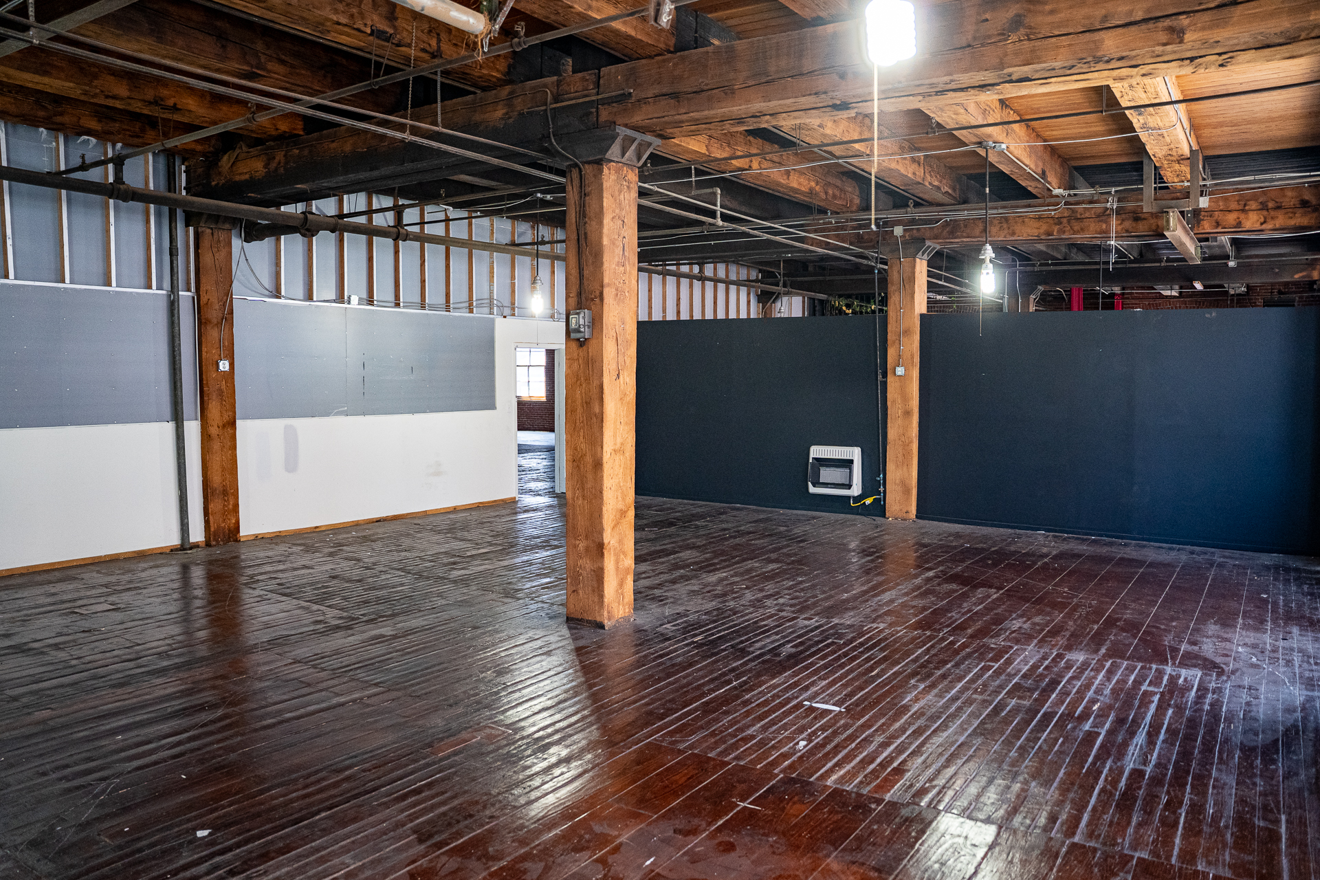 A wide shot of an empty, large industrial space with a shiny, dark wooden floor and a ceiling crisscrossed with exposed wooden beams and pipes. Several thick wooden pillars are visible. The space is divided by temporary partition walls, with one section painted white and gray, and another solid black. A single, small window is visible in the background.