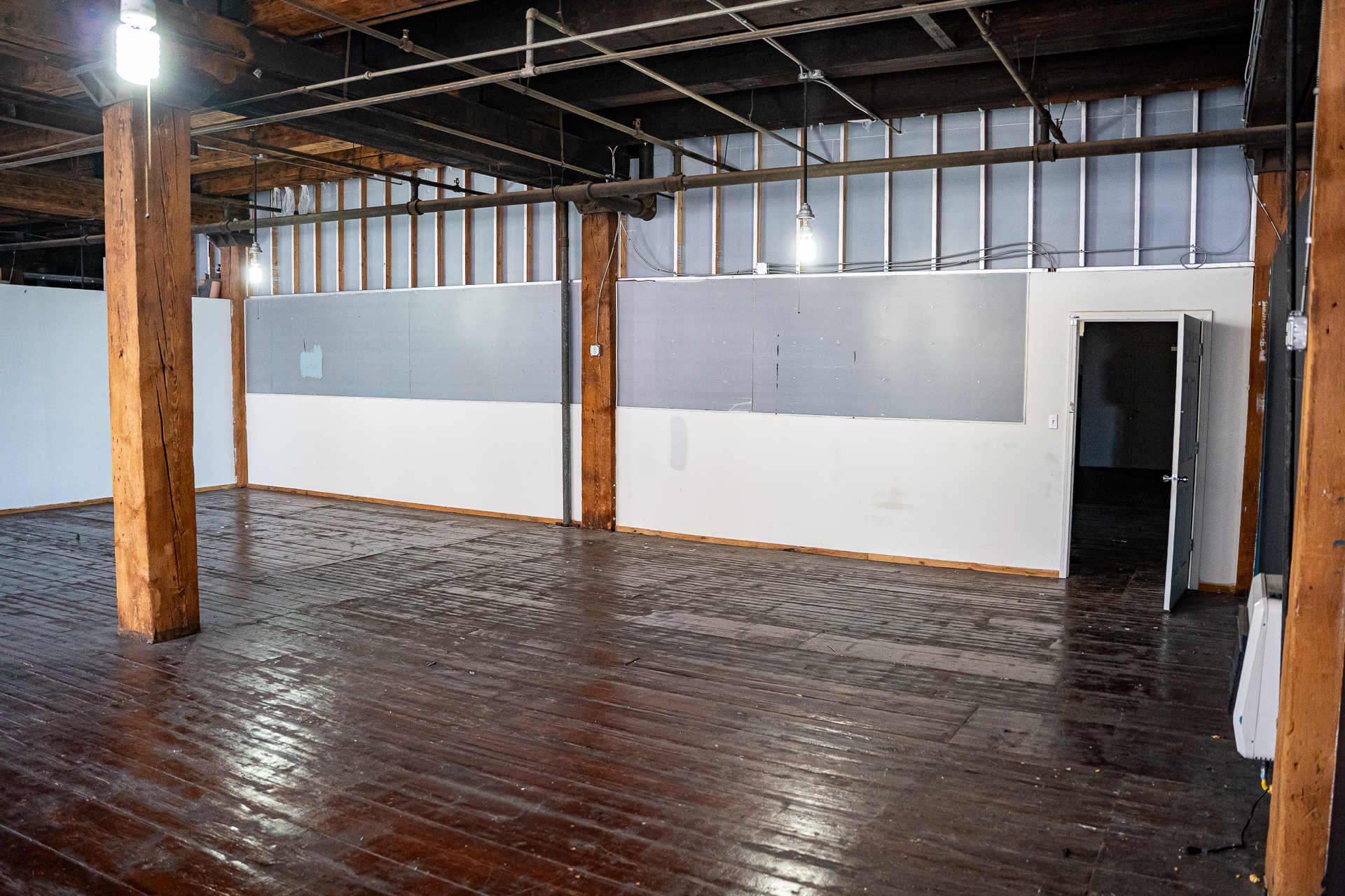 An empty industrial loft space with a dark, aged hardwood floor. Exposed wooden beams and pipes are visible on the ceiling. A partial wall, painted white on the bottom and gray on the top, runs across the width of the image. A wooden support pillar stands on the left. On the right, an open doorway leads to another part of the space. The floor shows signs of wear and tear.