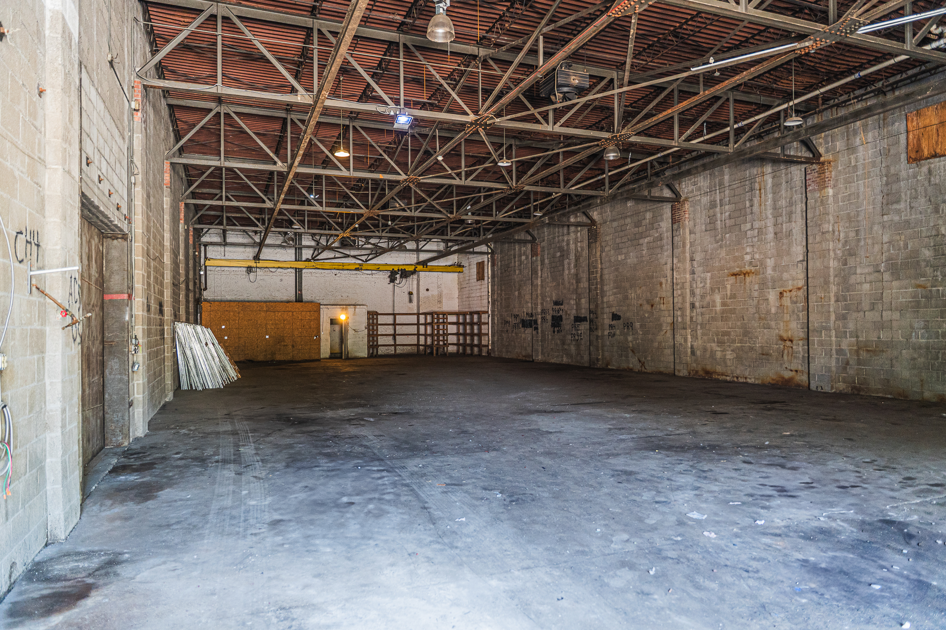 A wide-angle interior view of a large, empty industrial warehouse. The space has a rough concrete floor, and its walls are made of gray concrete blocks. The ceiling is high and features a grid of exposed metal truss beams. A yellow overhead crane rail is visible in the middle. A stack of long white materials leans against a wall on the left.