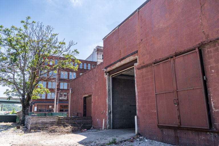 An exterior view of a weathered red industrial building. A large, open loading dock door is visible in the center, with a rusty, ribbed metal sliding door partially covering it. To the left of the loading dock is a smaller wooden door. A large, green-leaved tree stands in the foreground on the left, and a multi-story red brick building is visible behind it in the distance. The ground is a mix of concrete and gravel.
