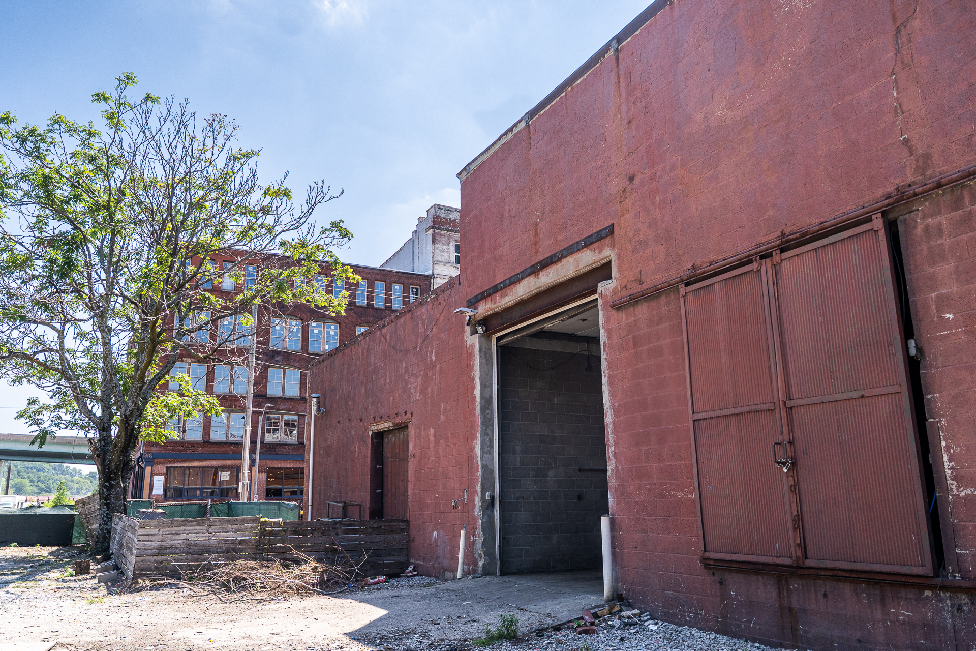 An exterior view of a weathered red industrial building. A large, open loading dock door is visible in the center, with a rusty, ribbed metal sliding door partially covering it. To the left of the loading dock is a smaller wooden door. A large, green-leaved tree stands in the foreground on the left, and a multi-story red brick building is visible behind it in the distance. The ground is a mix of concrete and gravel.