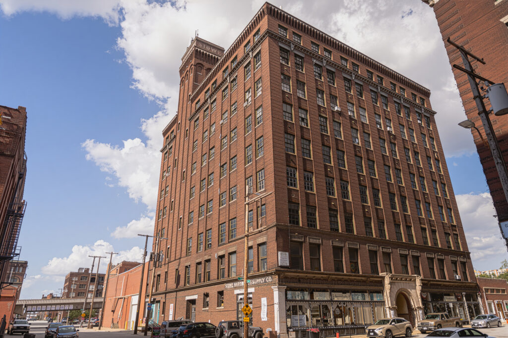 A low-angle, exterior view of a tall, historic red brick building, identified as the Stowe Building by a sign on its ground floor. The multi-story structure has many large windows and is situated on an urban street corner with other brick buildings visible. The sky is partly cloudy. Several cars are parked along the street in the foreground.